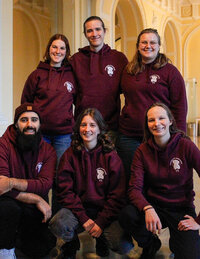 Foto: Eine Gruppe von Personen posiert in einem Innenraum. Sie tragen passende weinrote Kapuzenpullover mit einem Logo auf der Brust. Der Raum hat helle Wände und gewölbte Decken.