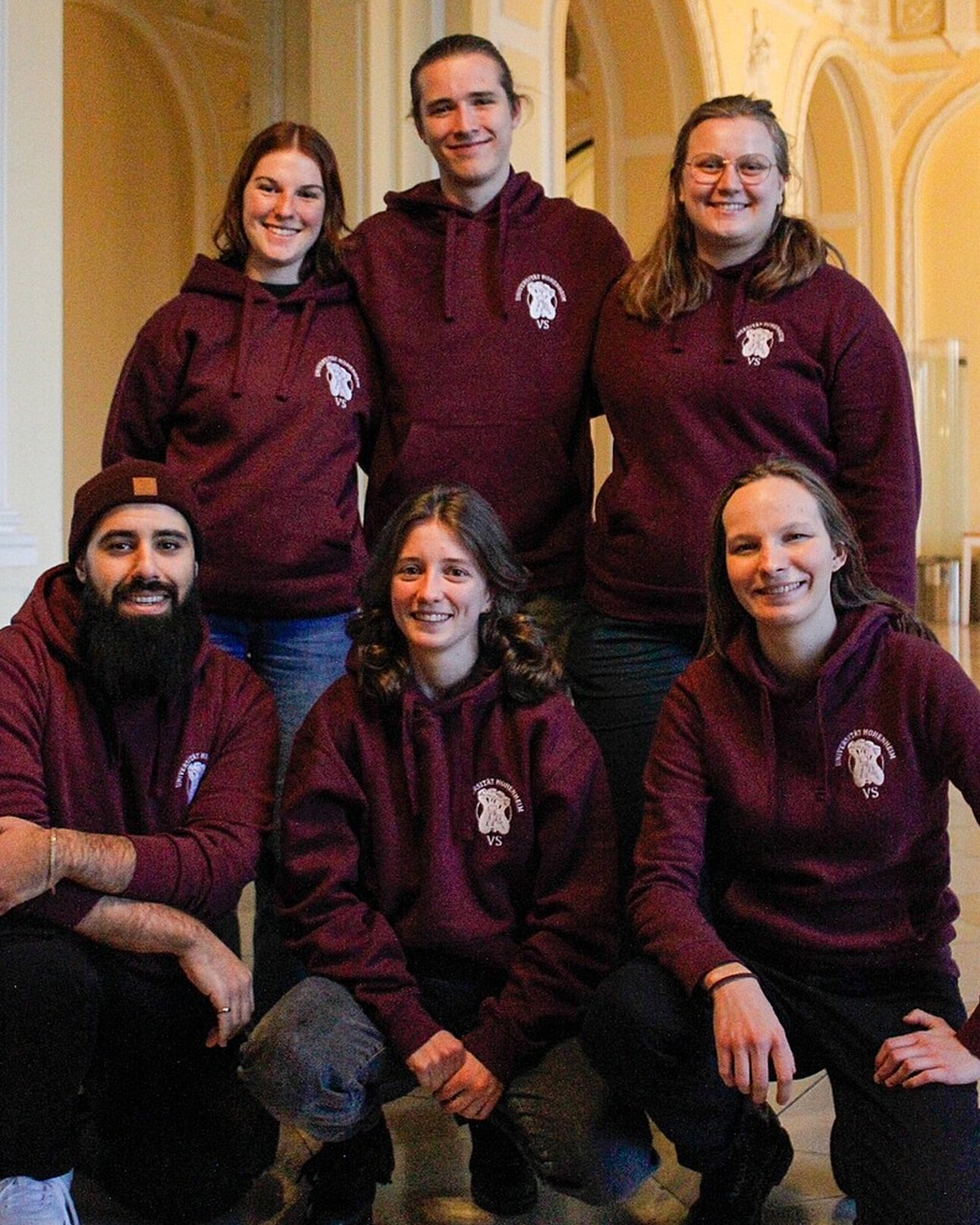 Foto: Eine Gruppe von Personen posiert in einem Innenraum. Sie tragen passende weinrote Kapuzenpullover mit einem Logo auf der Brust. Der Raum hat helle Wände und gewölbte Decken.