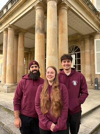 Foto: Drei Personen in bordeauxfarbenen Hoodies stehen vor den Säulen des Schloss Hohenheim. Die Architektur des Gebäudes ist klassisch mit großen Säulen und symmetrischem Design.