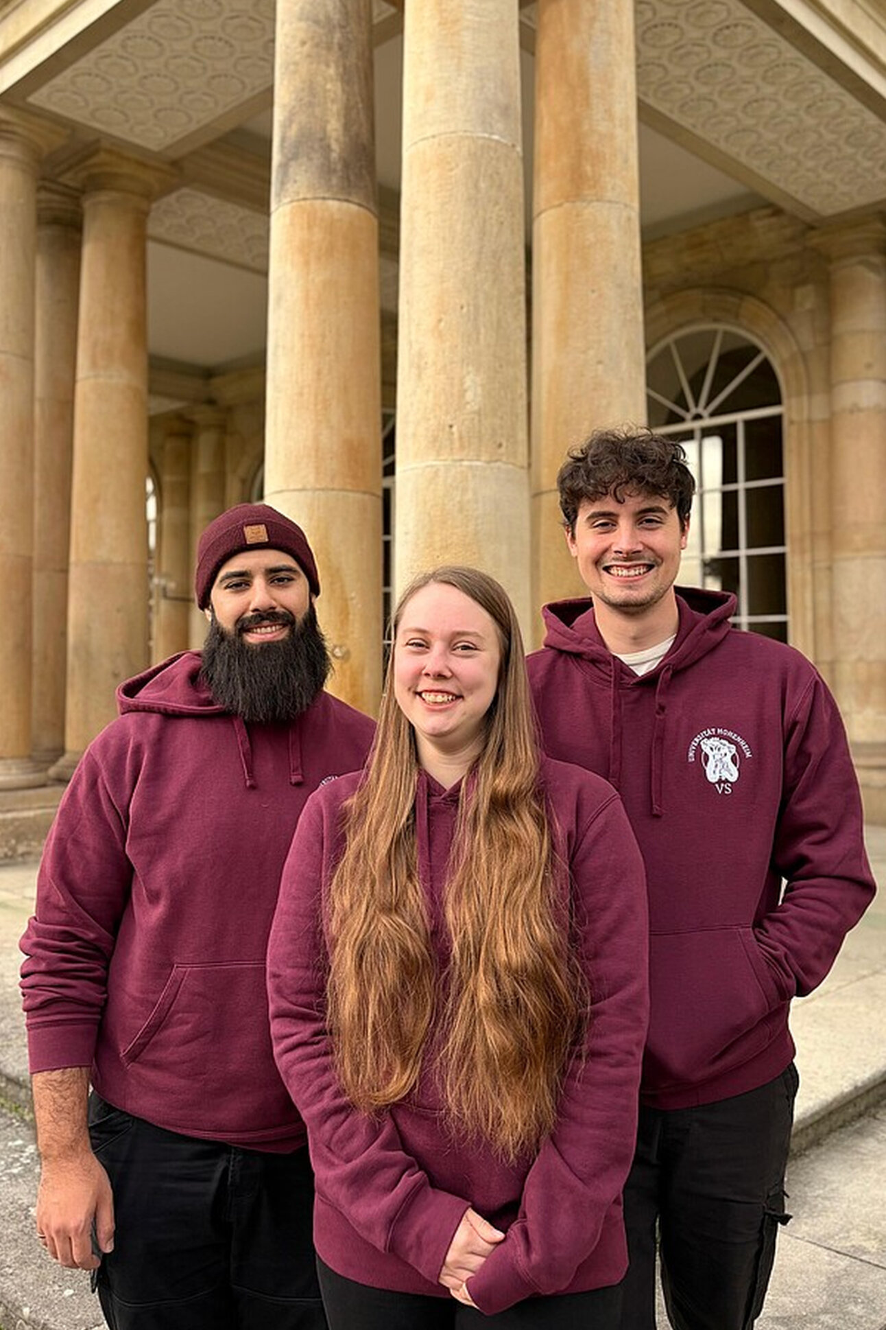 Foto: Drei Personen in bordeauxfarbenen Hoodies stehen vor den Säulen des Schloss Hohenheim. Die Architektur des Gebäudes ist klassisch mit großen Säulen und symmetrischem Design.