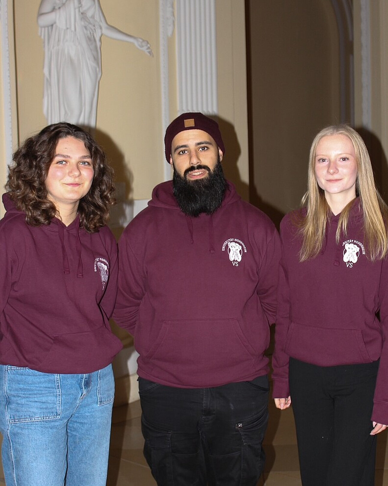 Foto: Drei Personen stehen nebeneinander in einem Innenraum vor einer Statue. Sie tragen passende bordeauxfarbene Hoodies mit einem Logo der Universität Hohenheim auf der Brust.