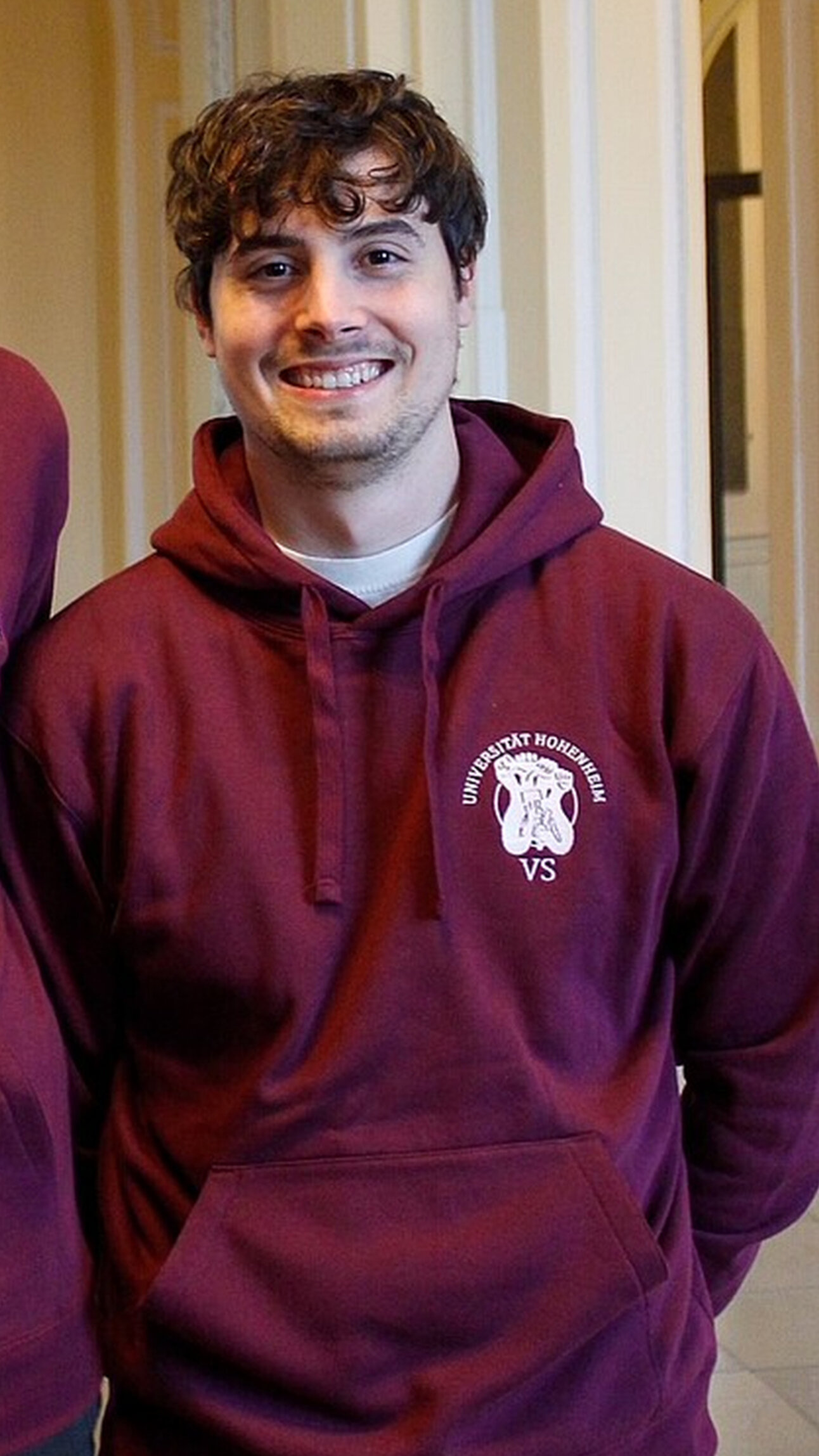 Foto: Zwei Personen tragen bordeauxfarbene Hoodies mit dem Logo der Universität Hohenheim. Sie stehen in einem Innenraum mit klassischer Architektur und Statuen.