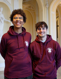 Foto: Zwei Personen tragen bordeauxfarbene Hoodies mit dem Logo der Universität Hohenheim. Sie stehen in einem Innenraum mit klassischer Architektur und Statuen.