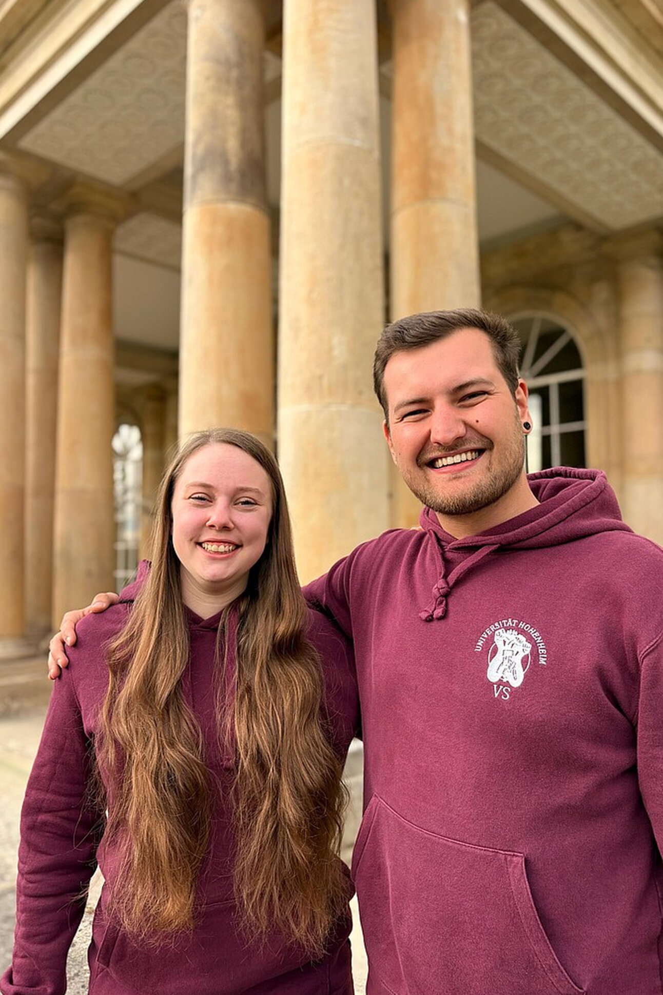 Foto: Zwei Personen in bordeauxfarbenen Hoodies mit dem Logo der Universität Hohenheim stehen vor den Säulen des Schloss Hohenheim.