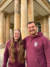 Foto: Zwei Personen in bordeauxfarbenen Hoodies mit dem Logo der Universität Hohenheim stehen vor den Säulen des Schloss Hohenheim.