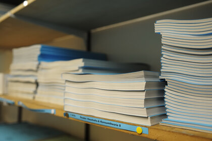 Foto: Mehrere Stapel von Büchern liegen auf einem Regal. Ein Etikett auf dem Regalbrett trägt die Aufschrift 'Konsumtheorie II'.
