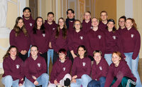 Foto: Eine Gruppe von Personen trägt bordeauxfarbene Kapuzenpullover mit einem Logo auf der Brust. Sie stehen und sitzen in einem Innenraum mit dekorativen Wänden.