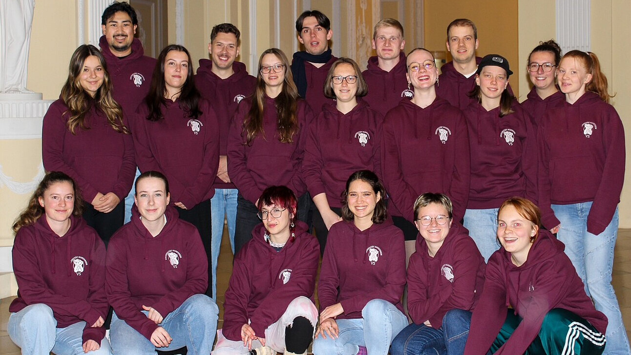 Foto: Eine Gruppe von Personen trägt bordeauxfarbene Kapuzenpullover mit einem Logo auf der Brust. Sie stehen und sitzen in einem Innenraum mit dekorativen Wänden.