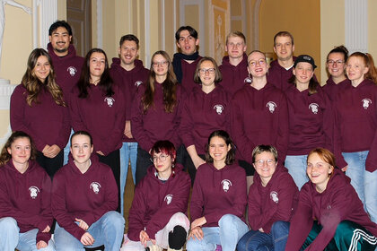 Foto: Eine Gruppe von Personen trägt bordeauxfarbene Kapuzenpullover mit einem Logo auf der Brust. Sie stehen und sitzen in einem Innenraum mit dekorativen Wänden.