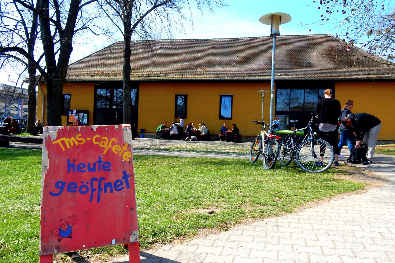 Foto: Ein rotes Schild mit der Aufschrift 'TMS-cafete heute geöffnet' steht auf einer Wiese. Im Hintergrund sind ein gelbes Gebäude, Fahrräder und Personen zu sehen.