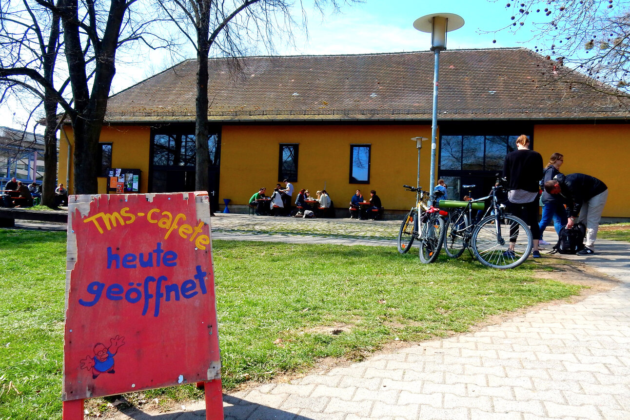 Foto: Ein rotes Schild mit der Aufschrift 'TMS-cafete heute geöffnet' steht auf einer Wiese. Im Hintergrund sind ein gelbes Gebäude, Fahrräder und Personen zu sehen.