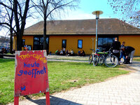Foto: Ein rotes Schild mit der Aufschrift 'TMS-cafete heute geöffnet' steht auf einer Wiese. Im Hintergrund sind ein gelbes Gebäude, Fahrräder und Personen zu sehen.