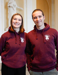 Foto: Zwei Personen tragen bordeauxfarbene Hoodies mit dem Logo der Universität Hohenheim. Sie stehen in einem Innenraum mit einer Statue im Hintergrund.
