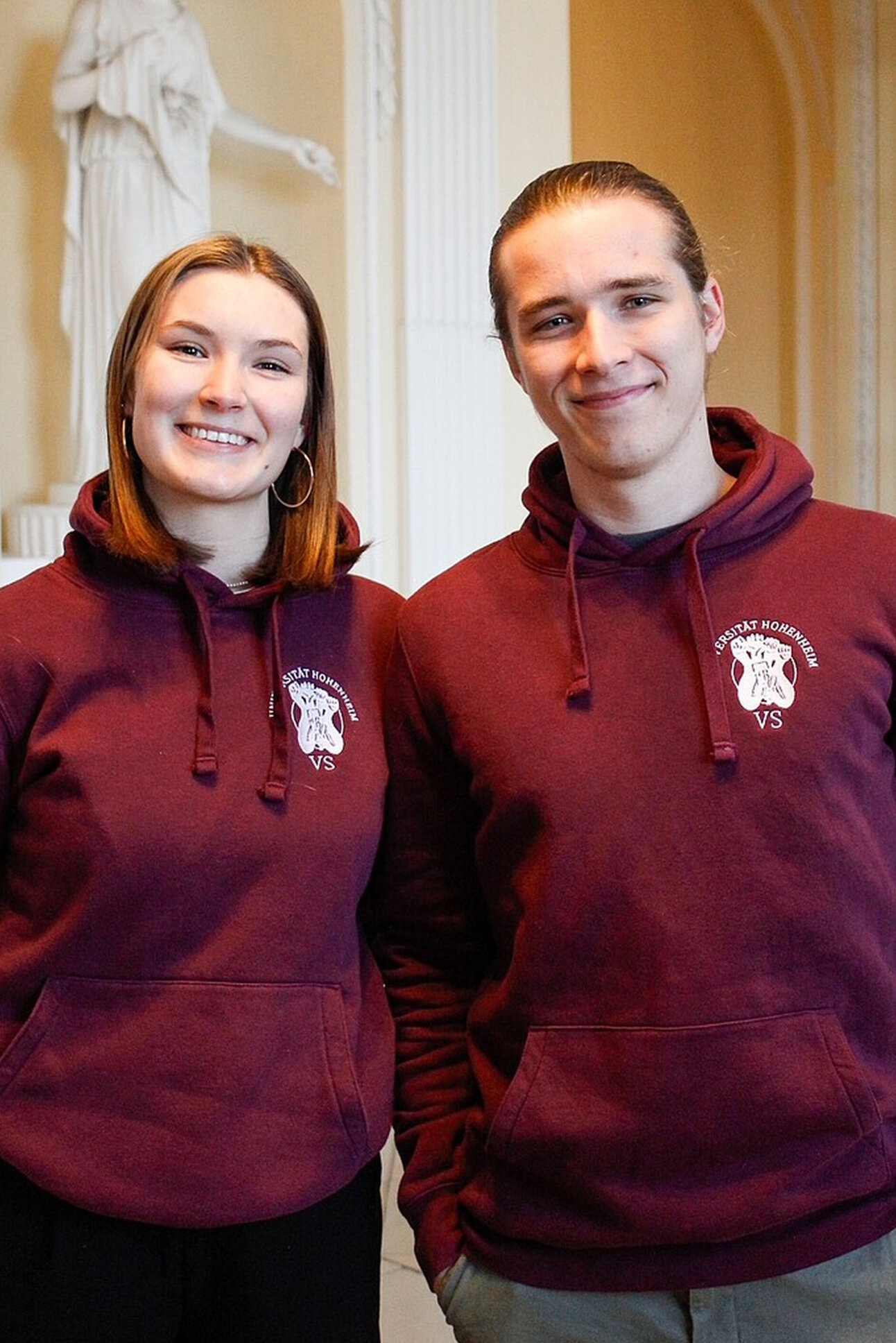 Foto: Zwei Personen tragen bordeauxfarbene Hoodies mit dem Logo der Universität Hohenheim. Sie stehen in einem Innenraum mit einer Statue im Hintergrund.