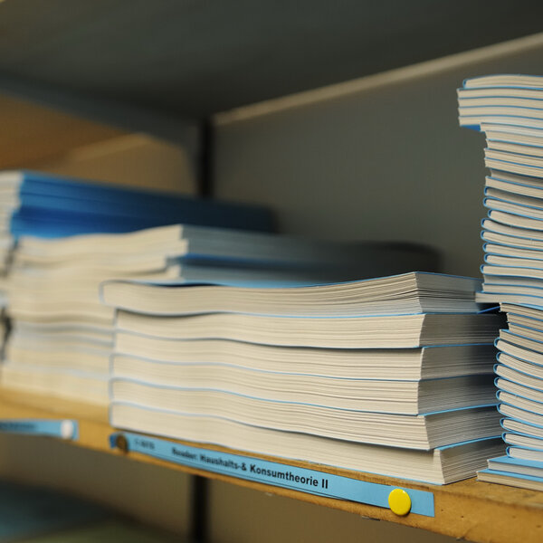 Foto: Mehrere Stapel von Büchern liegen auf einem Regal. Ein Etikett auf dem Regalbrett trägt die Aufschrift 'Konsumtheorie II'.