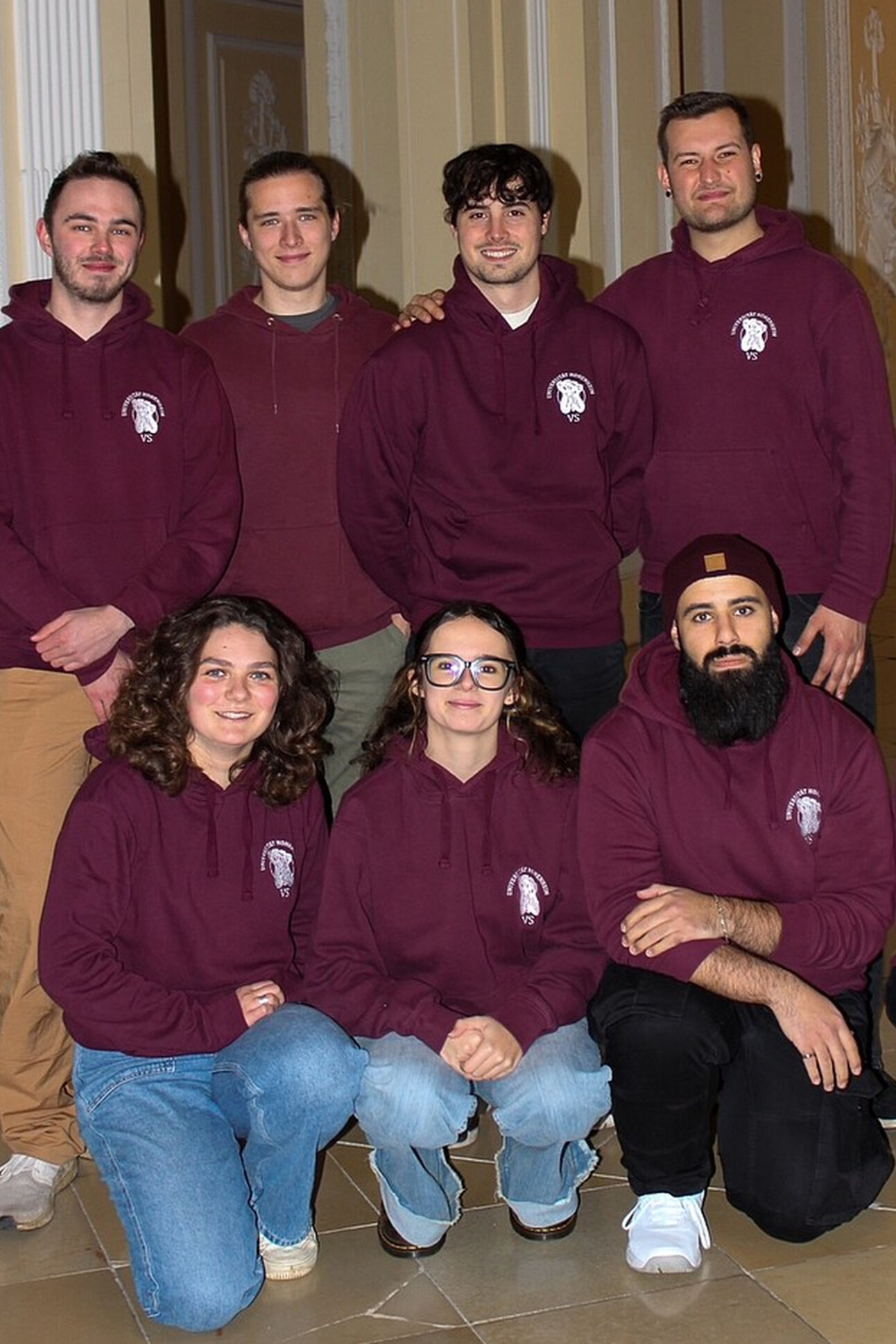 Foto: Eine Gruppe von Personen trägt bordeauxfarbene Kapuzenpullover mit einem Logo auf der Brust. Sie posieren in einem Innenraum mit dekorativen Wänden.