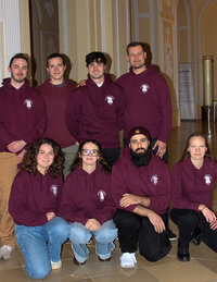 Foto: Eine Gruppe von Personen trägt bordeauxfarbene Kapuzenpullover mit einem Logo auf der Brust. Sie posieren in einem Innenraum mit dekorativen Wänden.