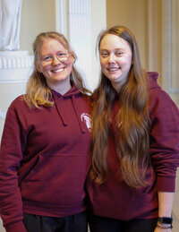 Foto: Zwei Personen tragen bordeauxfarbene Hoodies mit einem Logo der Universität Hohenheim. Sie stehen in einem Innenraum mit hellen Wänden und dekorativen Elementen.