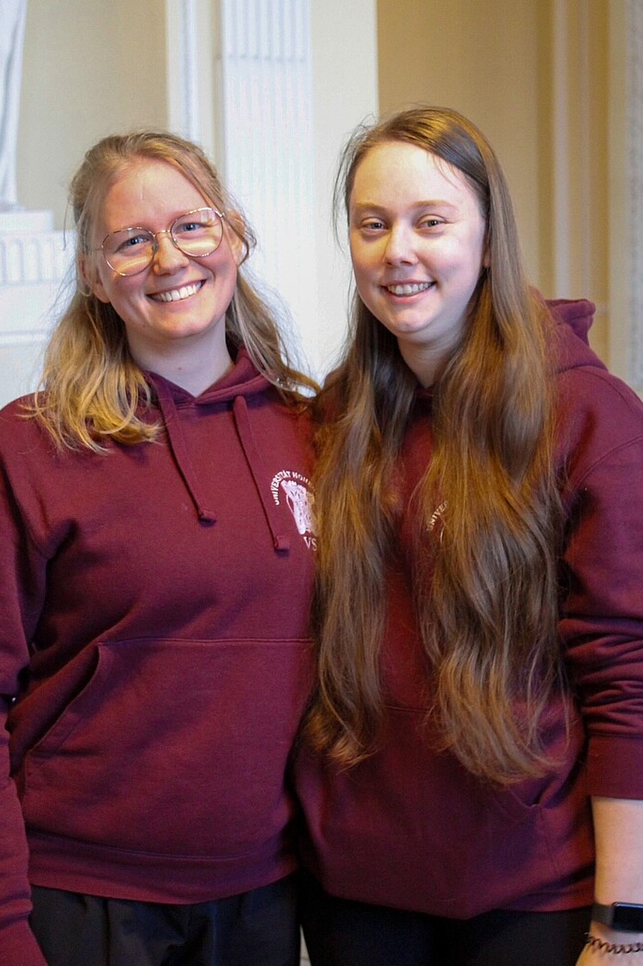 Foto: Zwei Personen tragen bordeauxfarbene Hoodies mit einem Logo der Universität Hohenheim. Sie stehen in einem Innenraum mit hellen Wänden und dekorativen Elementen.