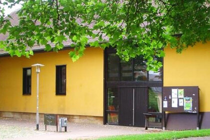 Foto: Ein gelbes Gebäude mit großen Fenstern und einem Schaukasten an der rechten Seite. Vor dem Gebäude stehen Bäume, die teilweise das Dach verdecken. Eine Laterne steht links im Bild.