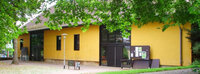 Foto: Ein gelbes Gebäude mit großen Fenstern und einem Schaukasten an der rechten Seite. Vor dem Gebäude stehen Bäume, die teilweise das Dach verdecken. Eine Laterne steht links im Bild.