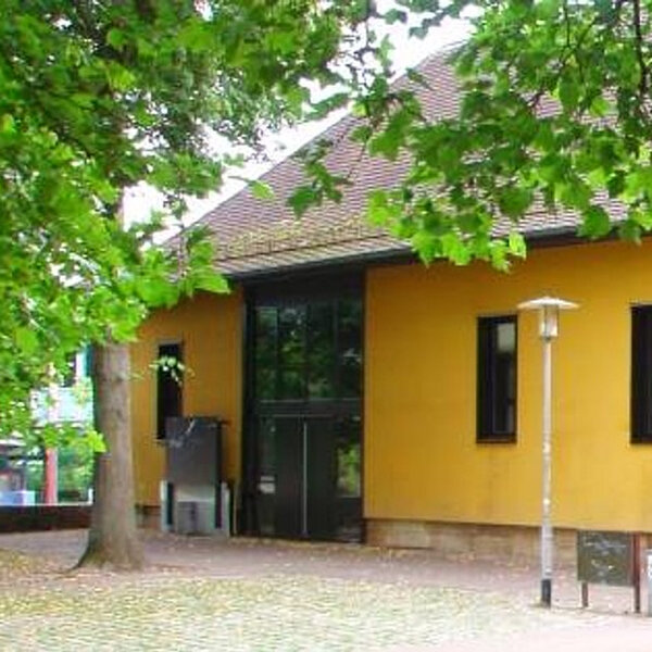 Foto: Ein gelbes Gebäude mit großen Fenstern und einem Schaukasten an der rechten Seite. Vor dem Gebäude stehen Bäume, die teilweise das Dach verdecken. Eine Laterne steht links im Bild.