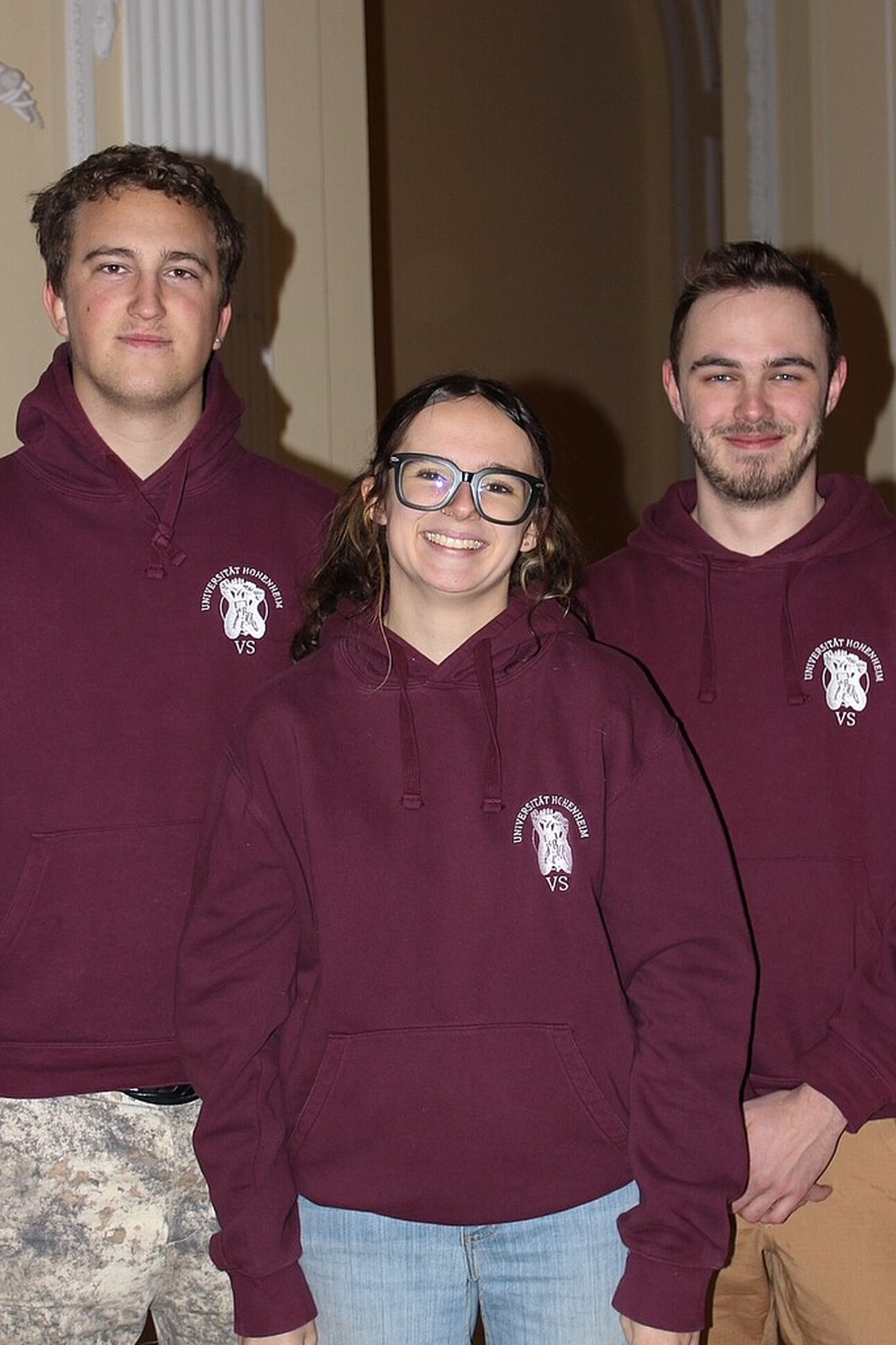 Foto: Drei Personen tragen bordeauxfarbene Hoodies mit dem Logo der Universität Hohenheim und der Aufschrift 'VS'. Sie stehen vor einem neutralen Hintergrund in einem Innenraum.