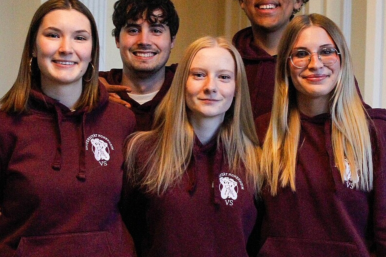 Foto: Fünf Personen stehen in einem Innenraum und tragen bordeauxfarbene Hoodies mit dem Logo der Universität Hohenheim. Die Personen stehen eng beieinander vor einem hellen Hintergrund mit Säulen.