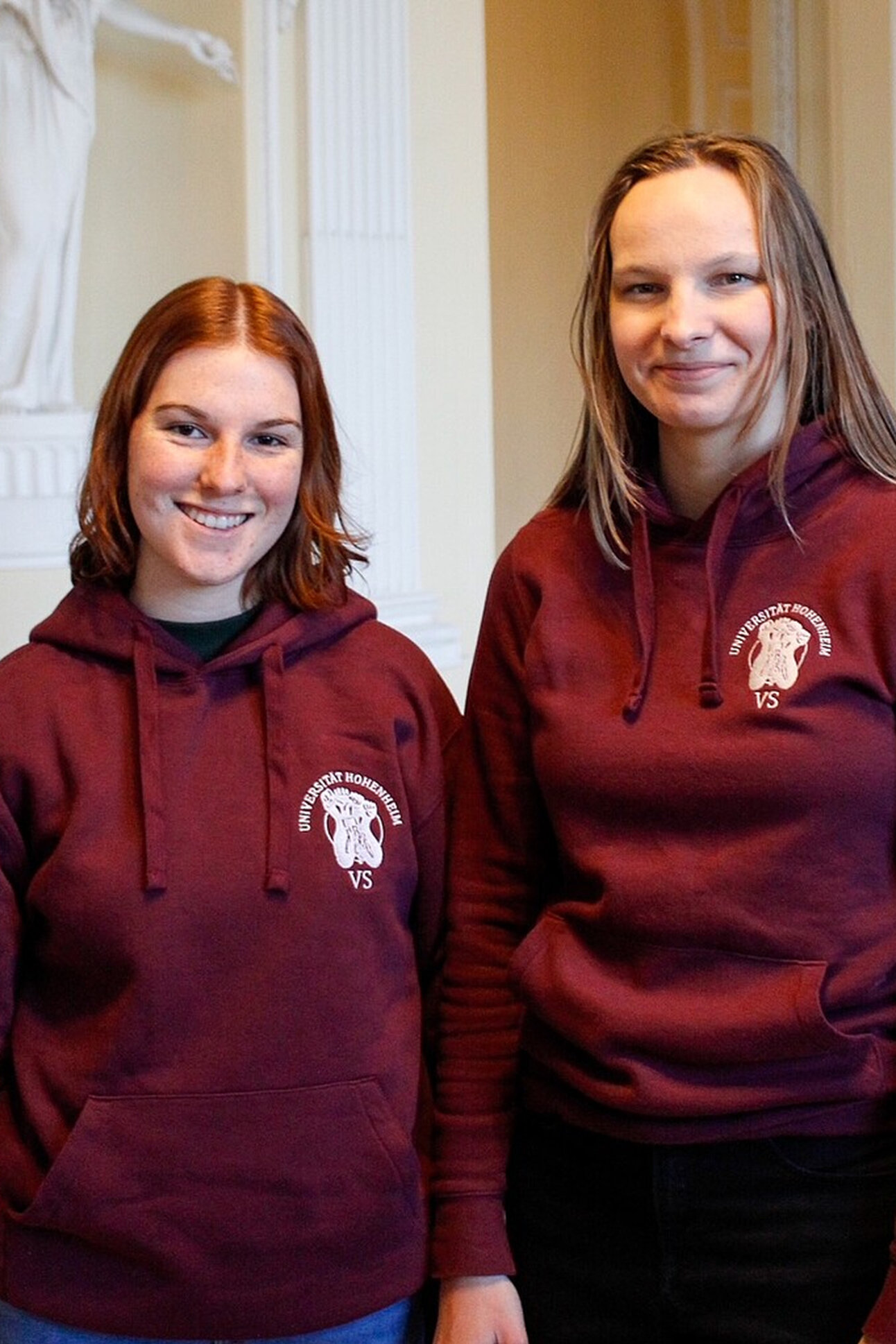 Foto: Zwei Personen tragen bordeauxfarbene Hoodies mit dem Logo der Universität Hohenheim. Sie stehen in einem Innenraum mit dekorativen Säulen im Hintergrund.