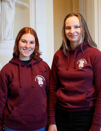 Foto: Zwei Personen tragen bordeauxfarbene Hoodies mit dem Logo der Universität Hohenheim. Sie stehen in einem Innenraum mit dekorativen Säulen im Hintergrund.