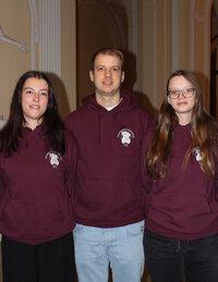 Foto: Drei Personen stehen nebeneinander in einem Innenraum. Sie tragen bordeauxfarbene Hoodies mit dem Logo der Universität Hohenheim und der Aufschrift 'VS'.