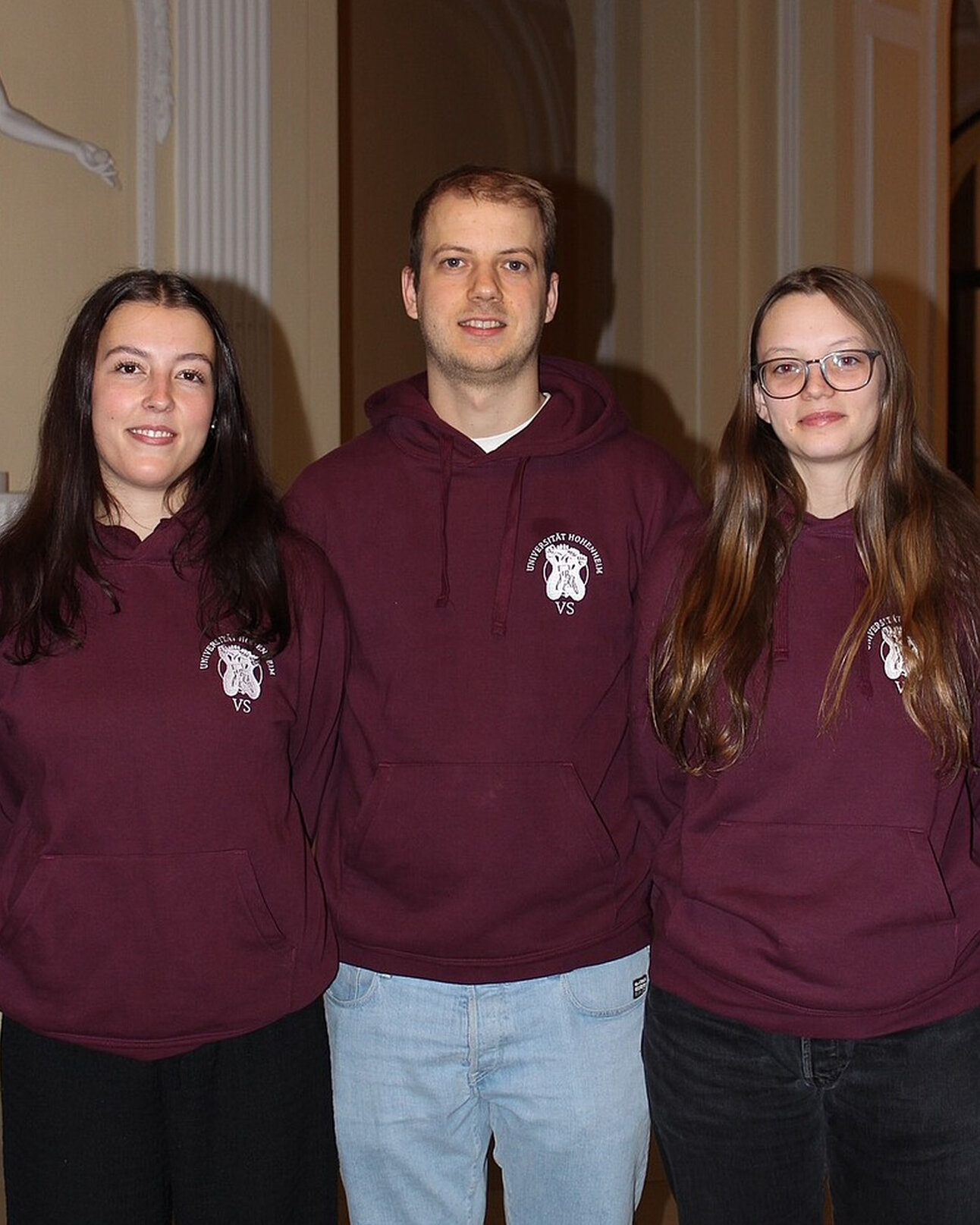 Foto: Drei Personen stehen nebeneinander in einem Innenraum. Sie tragen bordeauxfarbene Hoodies mit dem Logo der Universität Hohenheim und der Aufschrift 'VS'.