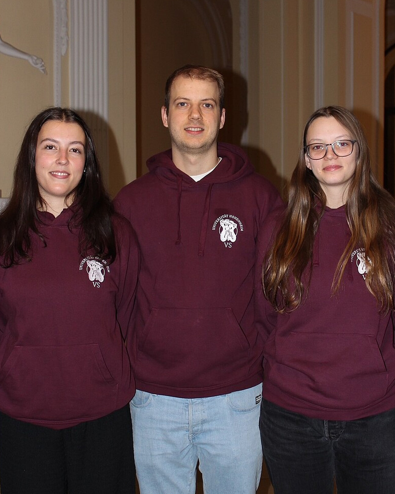 Foto: Drei Personen stehen nebeneinander in einem Innenraum. Sie tragen bordeauxfarbene Hoodies mit dem Logo der Universität Hohenheim und der Aufschrift 'VS'.