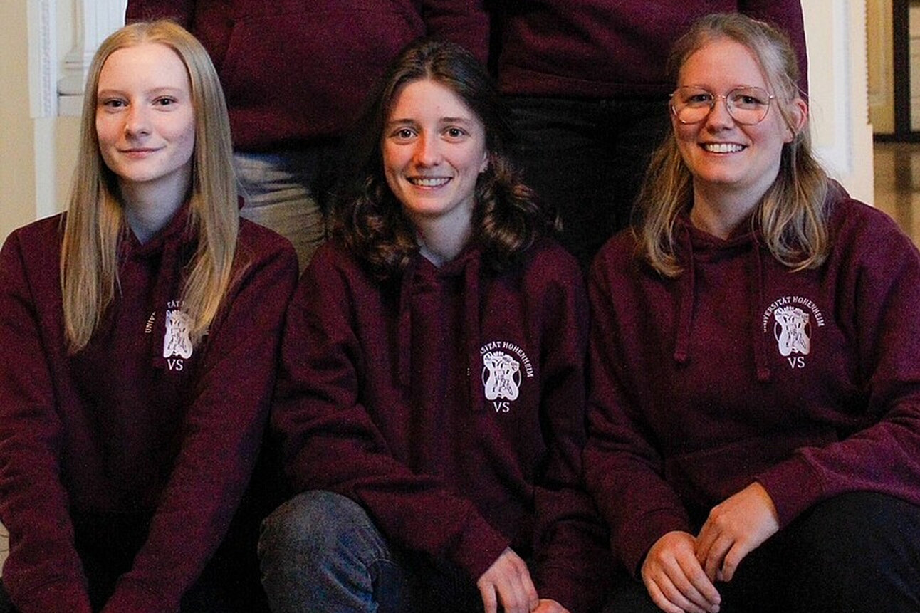 Foto: Fünf Personen stehen und sitzen in einem Innenraum. Sie tragen bordeauxfarbene Kapuzenpullover mit einem Logo auf der Brust. Der Hintergrund zeigt eine helle Wand mit architektonischen Details.